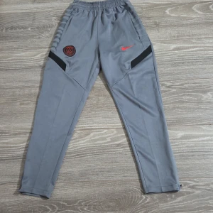 Grå Nike PSG träningsbyxor - Snygga grå träningsbyxor från Nike med PSG-logga och röd swoosh på framsidan. Byxorna har elastisk midja, svarta detaljer på låren och smal passform. Perfekta för fotbollsträning eller chill.