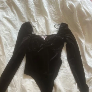 Svart sammetsbody med lång ärm - Supersnygg svart body  i sammet från Pieces med lång ärm och djup, något v-ringad halsringning. Kroppsnära passform och lätt puff vid axlarna ger en trendig siluett. Perfekt att styla med höga jeans eller kjol för en cool look.
