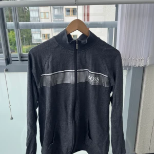 Mörkgrå zip hoodie från Hugo Boss - Mörkgrå zip hoodie från Boss med vita ränder över bröstet och logga på vänster sida. Jackan har hög krage, hel dragkedja och två fickor framtill. Perfekt för en chill och sportig look.