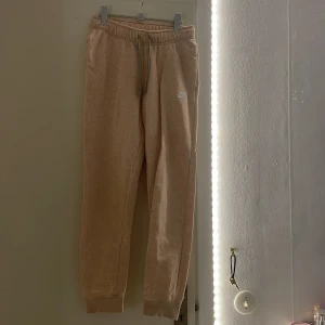 Beige mjukisbyxor från Nike - Säljer ett par beige mjukisbyxor från Nike med broderad logga på benet. Byxorna har elastisk midja med snörning och muddar vid bensluten. Perfekta för chill dagar eller när du vill vara extra bekväm. Dom va gråa innan men råka bleka dom så dom blev i denhär aprikos/beige färgen.