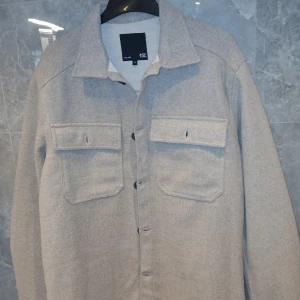 Grå overshirt från 157 med fickor - Snygg grå overshirt från 157 med klassisk krage och två stora bröstfickor med lock. Jackan är i mjukt material, har knäppning framtill och långa ärmar med knapp vid ärmslut. Perfekt för lager-på-lager och chill stil.