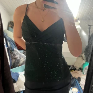 Glittrig svart-grön linnetopp Orsay - Snygg linnetopp från Orsay i svart med grönt glitter. Toppen har smala axelband, omlott-v-ringning och en liten lackdetalj med rosett under bysten. Perfekt för dig som vill sticka ut med lite extra bling. Materialet är mjukt och stretchigt syntet.