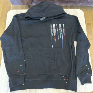Svart Amiri hoodie med färgstänk - Svart hoodie från Amiri med huva och känguruficka. Framsidan har AMIRI-logga med färg som rinner ner i vitt, blått och rött. Färgstänk i blått, rött och gult på ärmar och nedtill ger en kreativ vibe. Perfekt för dig som gillar streetwear med edge. Dm för storlekar 