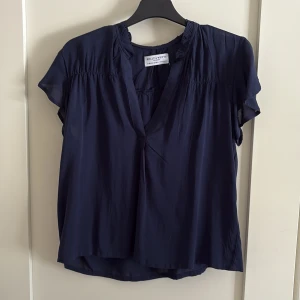 Mörkblå blus från Holly & Whyte XS - Mörkblå blus med korta volangärmar och v-ringning från Holly & Whyte by Lindex. Lätt och luftig känsla, rynkade detaljer vid axlar och halsringning. Perfekt för dig som gillar en enkel men stilren look.