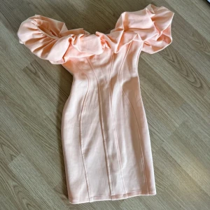 Ljusrosa kort klänning med volang - Superfin ljusrosa kortklänning med stora volanger över axlarna och offshoulder-modell. Figurnära passform som framhäver siluetten. 