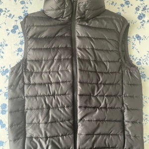 Svart dunväst från H&M, storlek S - Svart dunväst från H&M i storlek S. Västen är quiltad med horisontella sömmar, har hög krage och dragkedja framtill. Tillverkad i syntetmaterial och perfekt för lager-på-lager-stil. Enkel och clean design som funkar till många outfits.