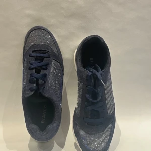 Blå glittriga sneakers från Bianco - Snygga mörkblå sneakers från Bianco med glittriga detaljer på ovandelen. Skorna har snörning, vit sula och en rund tå. Materialmix med textil och syntet ger en cool look som sticker ut.