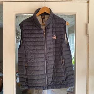 Mörkblå dunväst från Colmar - Snygg mörkblå dunväst från Colmar med quiltad design och dragkedja framtill. Västen har två fickor med dragkedja och en hög krage. Perfekt att slänga över en hoodie eller tröja för extra värme. Colmar-logga på bröstet ger en sportig vibe.