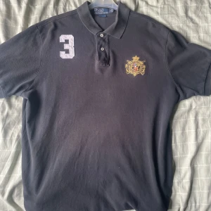 Svart pikétröja Polo Ralph Lauren  - Klassisk svart pikétröja från Polo Ralph Lauren i 100% bomull. Tröjan har broderad logga på bröstet, vit siffra 3 på axeln och snygg krage med knappar. Custom fit-modell som passar perfekt till en stilren look.