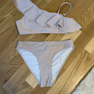 Randig bikini med volang och asymmetrisk axel - Superfin bikini med smala beige och vita ränder. Toppen har en axel och volangdetalj som ger en trendig look. Bikinin är fodrad och har en klassisk bikiniunderdel. Perfekt för stranden eller poolen!