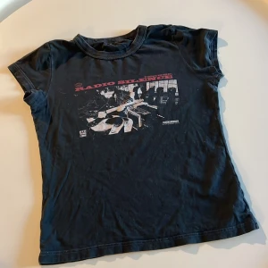 Svart t-shirt med Radio Silence-tryck - Cool svart t-shirt med tryck av en gitarrist och texten 'Radio Silence' i rött. Motiv med retro-vibe och New York 1989-detaljer. Klassisk rund hals och korta ärmar, perfekt för dig som gillar musik och vintage-stil.