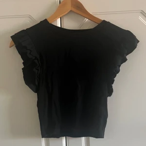Svart ribbad topp med volangärm Zara - Svart ribbad topp från Zara i storlek M. Toppen har snygga ärmar som ger en cool och trendig look. Materialet är stretchigt och sitter skönt på kroppen. Perfekt för dig som gillar stilrena plagg med en twist.