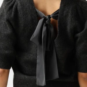 Ny Mörkgrå stickad topp med rosett Neo Noir, strl 38 - Supersnygg mörkgrå stickad topp från Neo Noir med korta puffärmar och en stor rosett i ryggen. Modellen är croppad och har en öppen rygg med knyt. Perfekt för dig som vill ha något unikt och trendigt.