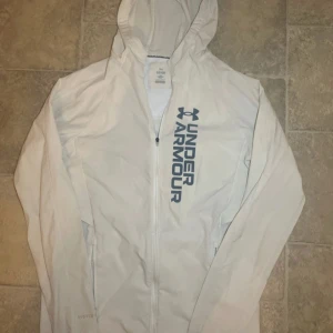 Vit vindjacka från Under Armour - Snygg vit vindjacka från Under Armour med huva och dragkedja framtill. Jackan har stora loggan tryckt på bröstet och meshfoder på insidan för extra komfort. Perfekt för träning eller när du vill ha en sportig look. Fickor med dragkedja på sidorna.