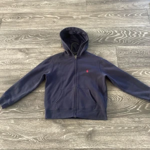 Marinblå hoodie från Polo Ralph Lauren - Snygg marinblå hoodie från Polo Ralph Lauren med dragkedja och klassisk röd broderad logga på bröstet. Tröjan har huva, kängurufickor och är tillverkad i mjuk bomull. Perfekt för en chill och stilren look.