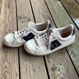 Vita sneakers från Fred Perry - Snygga vita sneakers från Fred Perry med marinblå detaljer och guldigt broderat lagerkransmärke på sidan och hälen. Skorna har klassisk snörning, rund tå och är tillverkade i skinn. Perfekta för en stilren och sportig look. 