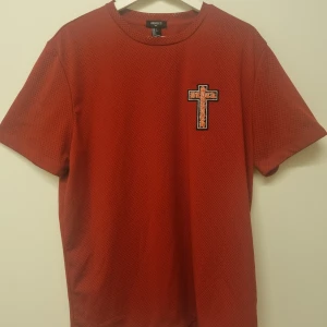 Röd mesh t-shirt - Röd mesh t-shirt med ett kors på bröstet. T-shirten har rund halsringning och korta ärmar.
