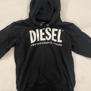Svart hoodie från Diesel med tryck - Svart hoodie från Diesel med stor vit logga och text på bröstet. Tröjan har huva med snörhål och är tillverkad i 100% bomull, vilket gör den både mjuk och skön. Perfekt för dig som gillar streetwear och vill sticka ut med en tydlig logga.