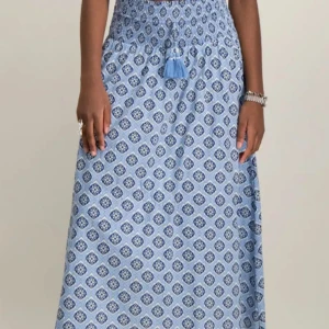 Blå långkjol med mönster från Gina Tricot - Supersöt långkjol från Gina Tricot i ljusblått med mörkblått och vitt mönster. Kjolen har smock i midjan, knytband med tofsar och ett flowigt fall. Perfekt för dig som gillar boho-vibe och vill ha något bekvämt och snyggt till sommaren.