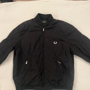 Svart bomberjacka från Fred Perry - Svart bomberjacka från Fred Perry med klassisk ribbad krage och muddar. Jackan har dragkedja framtill, två fickor med dragkedja och den ikoniska Fred Perry-loggan broderad på bröstet. Perfekt för dig som gillar stilren och sportig look.