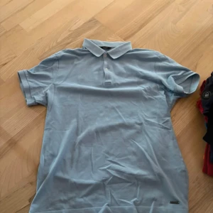 Ljusblå pikétröja Massimo Dutti - Snygg ljusblå pikétröja från Massimo Dutti i storlek S. Klassisk krage med två knappar och korta ärmar. Tillverkad i mjuk bomull som känns skön mot huden. Perfekt för en clean och stilren look.