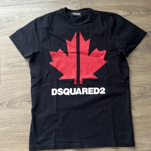 Svart DSQUARED2 t-shirt med lönnlöv - Svart t-shirt från DSQUARED2 med stort rött lönnlöv och vit logga på bröstet. Klassisk rund hals och korta ärmar. Materialet är mjuk bomull och trycket har små svarta prickar i lövet. Snygg och stilren design med sportig känsla.