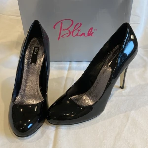 Svarta lackpumps från Blink, storlek 40 - Snygga svarta pumps från Blink i glansigt lackmaterial (patent PU). Klassisk modell med rund tå och 11 cm hög klack. Guldiga detaljer vid hälen och stilren design som ger en elegant look. Perfekta för dig som vill sticka ut med stil. Nya i kartong. Litet märke på ena hälen, fanns vid köp.