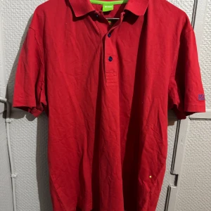 Röd pikétröja från Hugo Boss - Röd pikétröja från Hugo Boss med blå broderad logga på ärmen. Klassisk krage, knappar framtill och regular fit. Tillverkad i mjuk bomull och perfekt för en clean och stilren look.