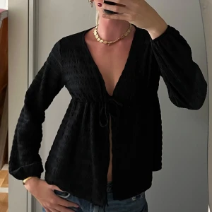 Svart blus med knyt och v-ringning - Svart blus med djup v-ringning och knyt framtill. Långärmad modell i ett lätt, strukturerat tyg som ger en luftig känsla. Blusen har en lös passform och ballongärmar.