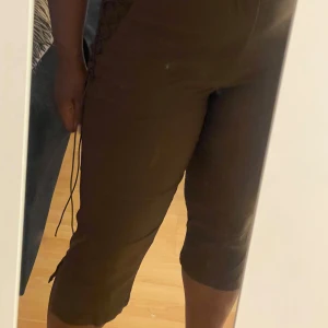 Bruna trekvartsbyxor med snörning - Säljer ett par bruna trekvartsbyxor med snygg snörningsdetalj längs sidorna. Byxorna har en tight passform och är perfekta för dig som vill ha något unikt och bekvämt. Materialet känns mjukt och stretchigt, vilket gör dem sköna att bära hela dagen.