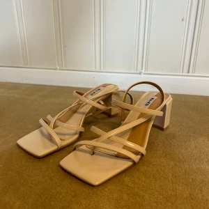 Beige klackar från NA-KD - Stilrena beige klackar  från NA-KD med fyrkantig tå, smala remmar och blockklack. Skorna har en modern design med öppen tå och är tillverkade i syntetmaterial.
