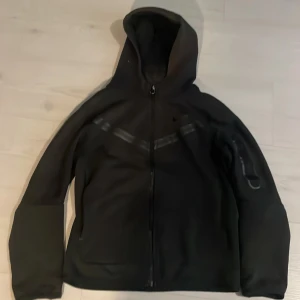 Svart hoodiejacka från Nike - Säljer en svart hoodiejacka från Nike med dragkedja framtill och huva. Jackan har långa ärmar och diskreta detaljer längs axlarna. Perfekt för en sportig och avslappnad stil.