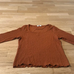 Rostbrun långärmad topp med volangkant - Superfin rostbrun långärmad topp med crinkle-textur och volangkant nertill och vid ärmslut. Modellen har en fyrkantig ringning och är stretchig och bekväm. Perfekt att styla med jeans eller kjol för en trendig look.