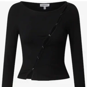 Svart ribbad topp  - Svart långärmad topp från Edited med ribbad struktur och en unik diagonal knappdetalj som skapar en cool öppning över bröstet. Figurnära passform och lätt croppad nertill. Perfekt för dig som gillar edgy och trendiga plagg. Storlek M men liten i storleken