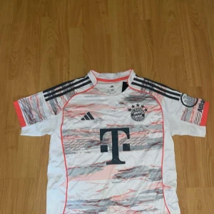Bayern München Adidas fotbollströja - Snygg Bayern München fotbollströja från Adidas med korta ärmar. Tröjan är vit med grå, rosa och svarta detaljer samt ett digitalt mönster över bröstet. Klassiska Adidas-ränder på axlarna och klubbmärke på bröstet. Perfekt för fans!