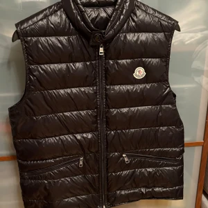 Moncler Gui Gilet/Vest - Moncler Gui väst i svart, storlek 3 (motsvarar M). Klassisk lättviktsväst med diskret glans, dunfyllning och ikoniska loggan på bröstet.  I nyskick – endast använd ett fåtal gånger. Inga defekter eller märken (skick 10/10). Kvitto från originalköp medföljer.  Säljer då den inte längre används.