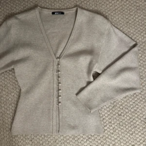 Beige kofta från Gina Tricot med knappar - Superfin beige/ grå kofta från Gina Tricot med v-ringning och klädda knappar framtill. Mjuk och skön, perfekt för lager-på-lager. Långärmad och i en stilren, enkel design som funkar till allt. Materialet känns som bomull eller en bomullsblandning. Orginalpris 399