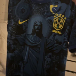 Blå Brasilien fotbollströja Nike - Snygg blå Brasilien fotbollströja från Nike med unikt tryck av Kristusstatyn och detaljer i gult. Tröjan har korta ärmar, Brasilien-emblem med stjärnor och Nike-logga på bröstet. Perfekt för dig som vill sticka ut på match eller träning.