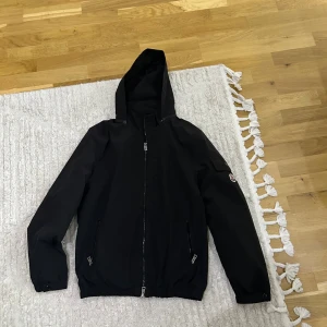 Svart vindjacka från Moncler med huva - Svart vindjacka från Moncler med huva, dragkedja framtill och två fickor med dragkedja. Jackan har en patch med Moncler-logga på ärmen och är tillverkad i ett lätt syntetmaterial. Perfekt för dig som gillar stilren och sportig look. OBSERVERA: köpt av en annan person på plick, fick inte med certifiering eller kvitto på att det är äkta. NFC funkar 