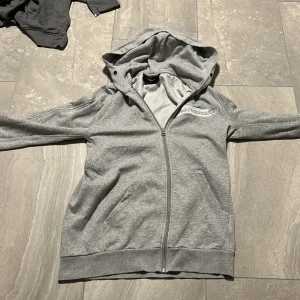 Grå hoodie från Peak Performance - Snygg grå hoodie från Peak Performance med dragkedja och broderad logga på bröstet. Tröjan har huva, två fickor framtill och är tillverkad i mjuk bomullsmix. Perfekt för chill dagar eller när du vill ha en clean och sportig look.