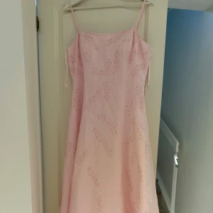 Ljusrosa långklänning med broderi - Superfin ljusrosa långklänning med tunna axelband och broderade detaljer i form av blad. Klänningen har en elegant A-linjeform och dragkedja i ryggen. Perfekt för dig som vill ha en romantisk och söt look till speciella tillfällen.
