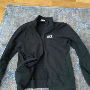 Svart EA7 zipjacka från Emporio Armani - Svart zipjacka från EA7 Emporio Armani med hög krage och vit logga på bröstet. Jackan har långa ärmar, dragkedja framtill och diskreta vita ränder på ryggen. Tillverkad i mjuk bomull, perfekt för en clean och sportig look.