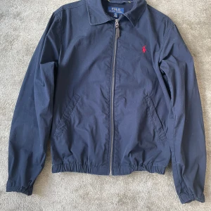 Marinblå vindjacka från Polo Ralph Lauren - Säljer sonens marinblåa vindjacka från Polo Ralph Lauren i 100% bomull. Klassisk design med dragkedja, två snedställda fickor och elastisk midja. Ikoniska röda loggan broderad på bröstet. Perfekt för dig som gillar stilren och tidlös look.