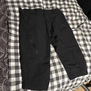 Svarta McKinley trekvartsshorts, storlek S - Svarta trekvartsshorts från McKinley i storlek S. Byxorna har trekvartslängd, elastisk midja och dragkedjeficka på ena benet. Perfekta för utomhusaktiviteter när du vill ha rörelsefrihet och skydd mot väder. Lätta och smidiga i syntetmaterial.