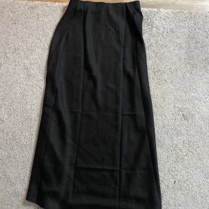 Svart långkjol i klassisk stil - En stilren svart långkjol med rak passform och enkel design. Kjolen är perfekt för dig som gillar minimalistiska plagg och funkar till många olika looks. Materialet känns mjukt och följsamt, och den höga midjan ger en snygg siluett.