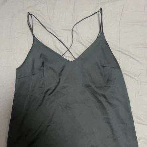 Svart topp med tunna axelband - Stilren svart topp med tunna axelband som korsas i ryggen. Toppen har v-ringad hals och ärmlös design, tillverkad i ett glansigt, mjukt material som ger en lyxig känsla. Perfekt för dig som gillar minimalistisk och trendig stil.