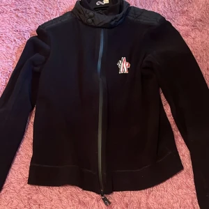 Svart Moncler jacka med dragkedja - Svart jacka från Moncler med hög krage och hel dragkedja framtill. Jackan har ett broderat Moncler-märke på bröstet och quiltade detaljer på axlarna. Tillverkad i ett mjukt material som känns både sportigt och stilrent. Perfekt för dig som gillar clean och exklusiv look.