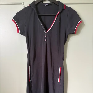Svart kort klänning Tommy Hilfiger storlek M  - Sportig svart kortklänning från Tommy Hilfiger med röda, vita och svarta detaljer vid krage, ärmslut och fickor. Klänningen har v-ringning med knappar och korta ärmar. Tillverkad i mjuk bomull, perfekt för en avslappnad och cool look. På mig som är 161 cm lång går den till en bit ovan knäna. Passar byst upp till 98 cm, midja upptill 90 cm och höft till 110cm. 