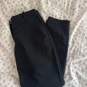 Svarta kostymbyxor från H&M - Klassiska svarta kostymbyxor från H&M i storlek 40. Byxorna har raka ben, hällor för bälte och stängs med hake och dragkedja. Perfekta för en clean och stilren look. Materialet känns mjukt och följsamt.