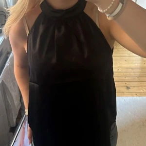 Svart halterneck topp - Snygg svart halterneck topp i glansigt satinliknande material. Toppen har hög hals med lätt rynk och är ärmlös med öppen rygg. Perfekt för dig som gillar stilrena och trendiga plagg med en elegant touch. Köpt för 70kr och endast provad